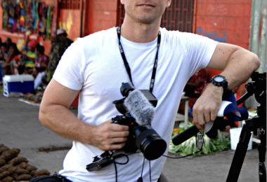 A young male journalist in a white tshirt with a camera around his neck leans against a tripod in a village street.
